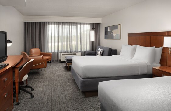 Typical Courtyard-style hotel guest room with carpet, upholstered seating, PTAC unit, drapes, hard surfaces, and multiple guest touchpoints